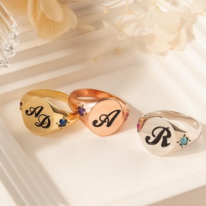 May include: Three signet rings with colorful gemstones. The rings are gold, rose gold, and silver. The rings are engraved with the letters "AD", "A", and "R".