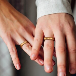 May include: Two gold rings with the words "JON" and "JOI" engraved on them. The rings are worn on the ring fingers of two hands.