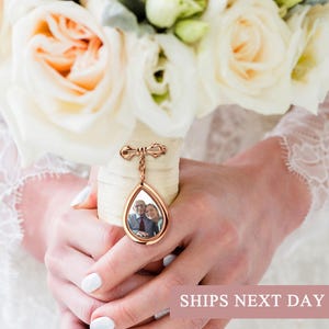 May include: A bouquet of white roses with a gold-colored teardrop-shaped locket. The locket contains a photograph and is attached to the bouquet with a decorative pin. The text "SHIPS NEXT DAY" is visible.