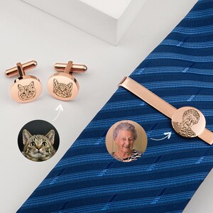 May include: A set of rose gold cufflinks and a tie bar with engraved images of a cat and a woman. The cufflinks and tie bar are on a blue patterned tie.