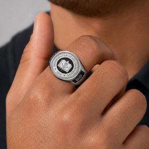 May include: A silver-toned signet ring featuring a circular black inlay with the text Brookhaven State University 1895 and an emblem of an open book with a torch. The side of the ring band displays the letters BS in a black rectangular field.
