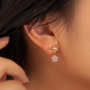 May include: A gold-toned earring featuring a small bow stud at the top with a dangling five-petaled flower charm. The flower charm is set with light pink stones and a small gold-toned center, creating a delicate and symmetrical jewelry piece.