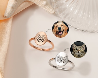 May include: Two silver rings with oval-shaped engravings of a dog and a cat. The rings are on a white surface with a seashell and a piece of fabric in the background.