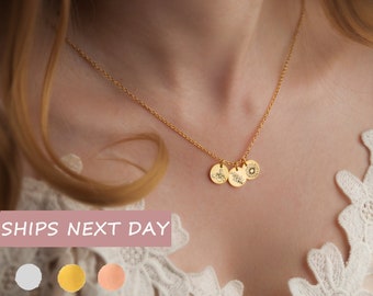 May include: A gold-colored necklace with three small, round pendants. Each pendant features a different engraved design: a bee, a sprig of leaves, and a sun. The necklace is displayed against a soft, neutral background.
