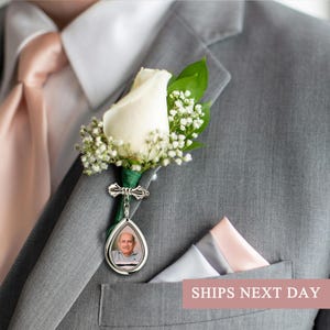 May include: A gray suit with a white rose boutonniere, baby's breath, and a silver pendant with a photo. A pink tie and pocket square complement the suit. The image shows a close-up of the lapel.