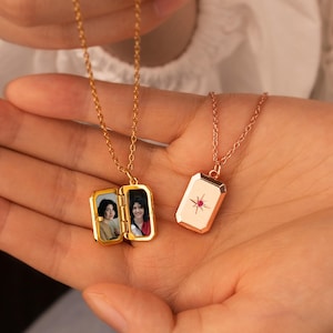 May include: Two gold-toned rectangular locket necklaces on delicate chains. One locket is open, revealing two small portrait photographs of a person. The other locket is closed, featuring a starburst engraving with a small red stone in the center.