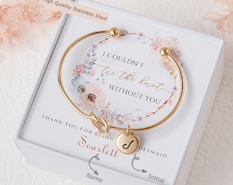 May include: Gold bracelet with a knot charm and a round charm with the letter J. The bracelet is in a white box with a floral design and the text "I couldn't tie the knot without you. Thank you for being Scarlett. Desmaid".