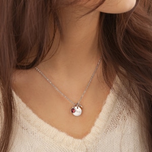 May include: A silver necklace with a small round pendant featuring the word "mom" in cursive script. A small red gemstone is attached to the pendant.