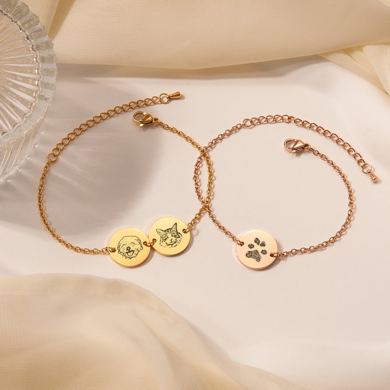 May include: Two gold chain bracelets with round charms. One bracelet has a charm with a dog's face and a cat's face. The other bracelet has a charm with a paw print.