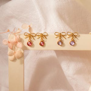 Puede incluir: Pendientes dorados con forma de lazo y gemas redondas colgantes. Dos pares presentan gemas rosas y los otros dos, moradas. Los pendientes se exhiben sobre una superficie crema contra un fondo blanco suave.