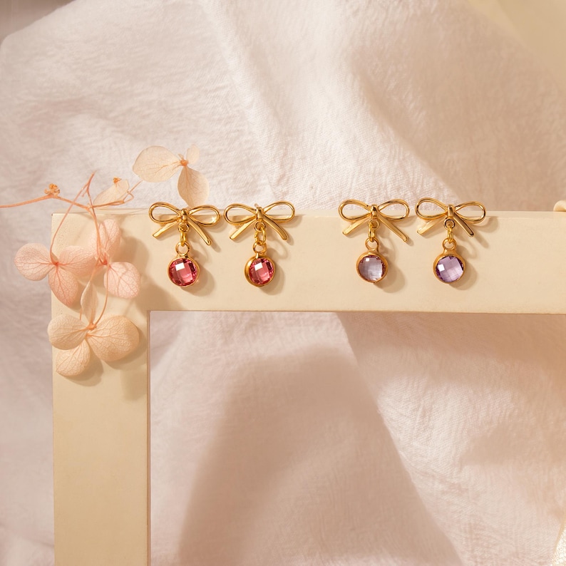 Puede incluir: Pendientes dorados con forma de lazo y gemas redondas colgantes. Dos pares presentan gemas rosas y los otros dos, moradas. Los pendientes se exhiben sobre una superficie crema contra un fondo blanco suave.