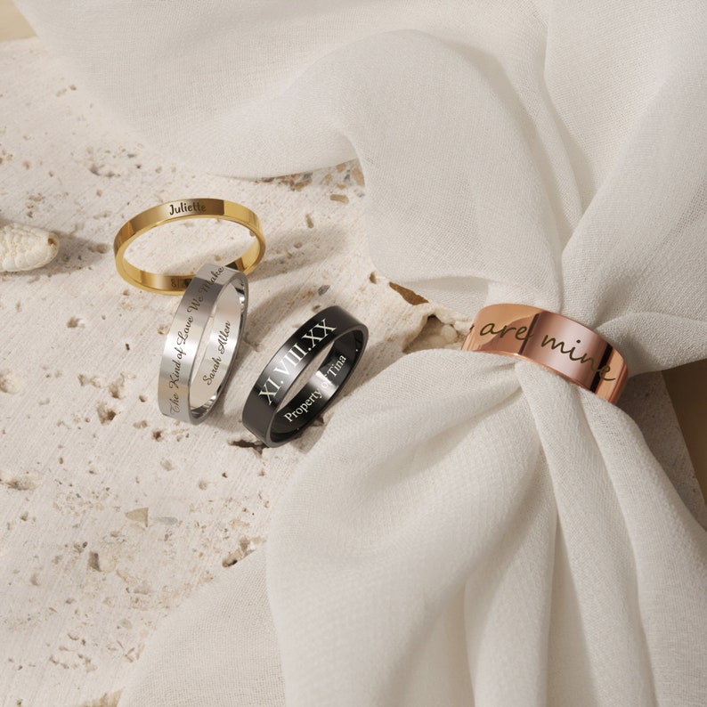 May include: Three engraved rings on a white fabric background. The gold ring says "Juliette". The silver ring says "The Kind of Love We Make Sarah Allan". The black ring says "XI.VIII.XX Property of Tina".