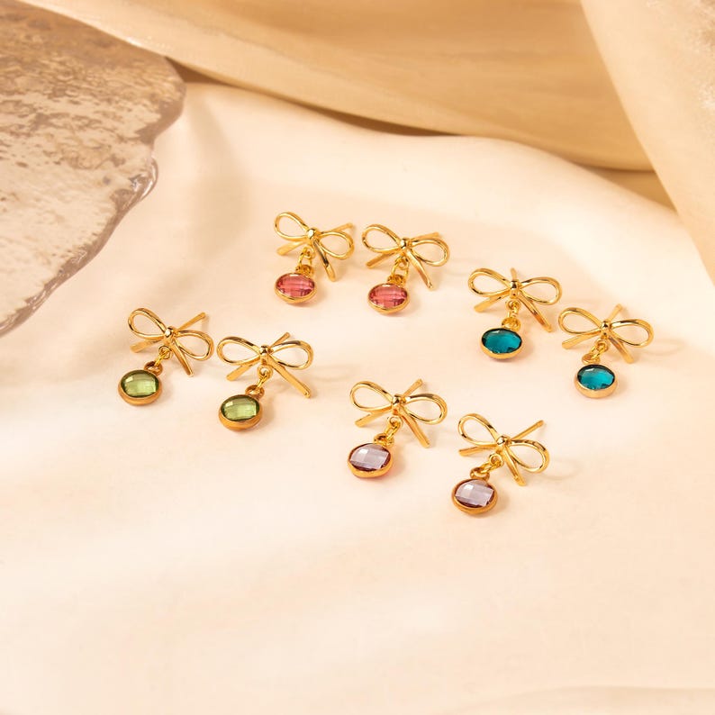 Puede incluir: Pendientes dorados con forma de lazo con gemas colgantes de colores. Los pendientes presentan un delicado dise&ntilde;o de lazo en la parte superior, con una variedad de colores de gemas que incluyen rosa, verde, azul y morado. Los pendientes se muestran sobre una superficie clara.