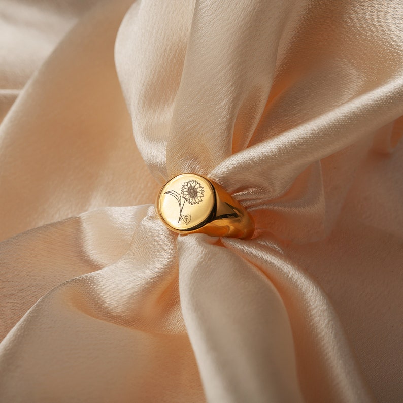 May include: A gold signet ring with a sunflower design engraved on the top. The ring is sitting on a soft, beige fabric.