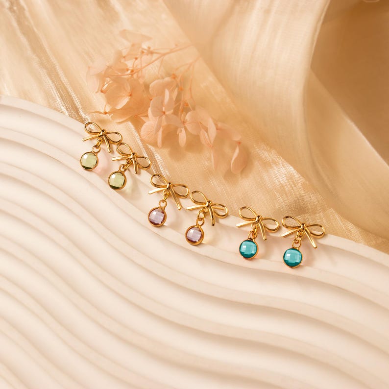 Puede incluir: Pendientes dorados con forma de lazo y gemas redondas de colores. Los pendientes presentan un delicado dise&ntilde;o de lazo y est&aacute;n adornados con piedras verdes, moradas y azules. Los pendientes se exhiben sobre una superficie color crema.
