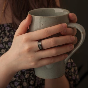 May include: A person's hand wearing a black ring with the inscription "unbreakable" on it. The hand is holding a light gray ceramic mug.