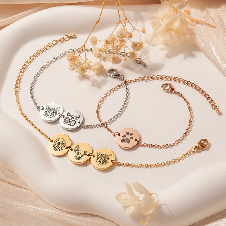 May include: Three gold, silver, and rose gold chain bracelets with charms. The gold bracelet has three round charms with engraved images of a dog, a cat, and a dog. The silver bracelet has two round charms with engraved images of a cat. The rose gold bracelet has one round charm with an engraved paw print.