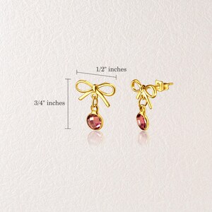 Puede incluir: Un par de pendientes dorados en forma de lazo con una gema rosa ovalada colgante. Los pendientes miden aproximadamente 1,9 cm de largo y 1,3 cm de ancho.