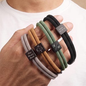 May include: Four double-cord bracelets in gray, tan, green, and black. Each features a square metal bead engraved with text: "Stay Hungry Hustle," "W/E/L/T," "K/W," and "Isaiah 43:18-19." The cords are woven, and the beads are dark metal.
