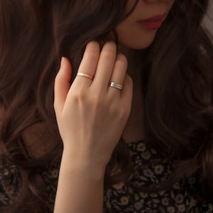 May include: A woman's hand wearing two rings. The ring on the index finger is a thin gold band. The ring on the middle finger is a silver band with the inscription "Always with you".