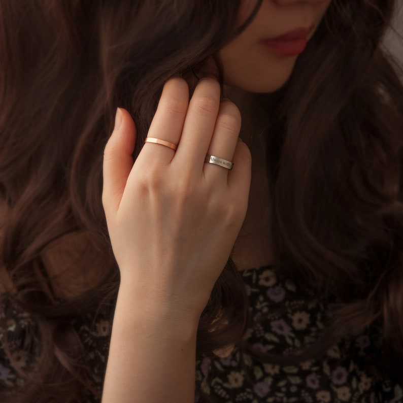 May include: A woman's hand wearing two rings. The ring on the index finger is a thin gold band. The ring on the middle finger is a silver band with the inscription "Always with you".