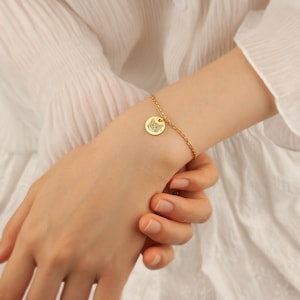 May include: Gold-colored bracelet featuring a small, round charm with a cat face design. The charm is attached to a delicate chain. The bracelet is worn on a wrist, showcasing the jewelry's design and color.