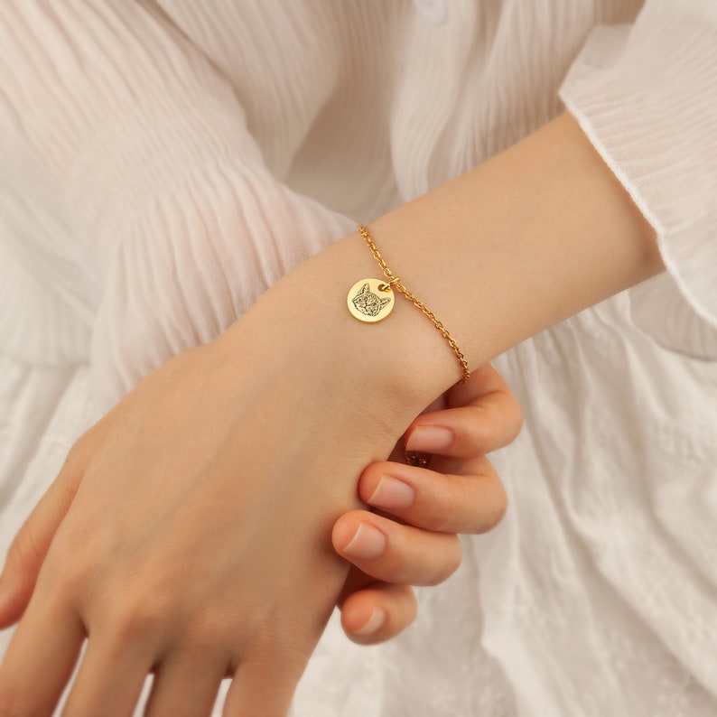 May include: Gold-colored bracelet featuring a small, round charm with a cat face design. The charm is attached to a delicate chain. The bracelet is worn on a wrist, showcasing the jewelry's design and color.