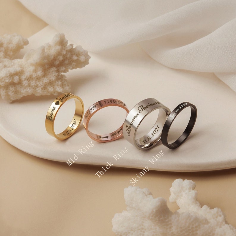 May include: Three personalized rings with different widths. The gold ring says "Laura Jade" and "3.25.2022". The rose gold ring says "4.6328&deg; N, 73.9524&deg; W" and "Always with you". The silver ring says "Brenna Theisen" and "11.8.2021".