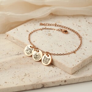 May include: Rose gold bracelet with three round charms, each engraved with a pet's face: a cat, a dog, and another cat. The bracelet is on a light-colored, textured surface.