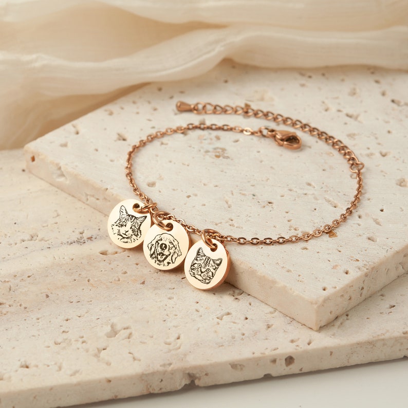 May include: Rose gold bracelet with three round charms, each engraved with a pet's face: a cat, a dog, and another cat. The bracelet is on a light-colored, textured surface.