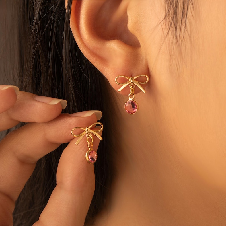 Puede incluir: Pendientes dorados con forma de lazo y una gema rosa colgante. Los pendientes se muestran en una oreja, con un segundo pendiente en la mano. El dise&ntilde;o de lazo a&ntilde;ade un toque delicado a la joya.