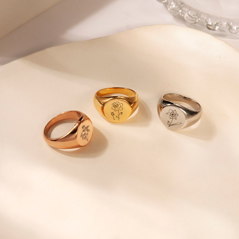 May include: Three gold signet rings with different floral designs. The rings are on a white surface.