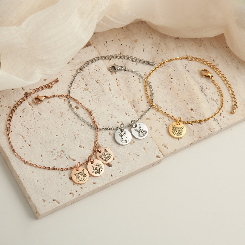May include: Three delicate bracelets in rose gold, silver, and gold. Each bracelet features a chain with a small, round pendant. The pendants are engraved with pet portraits, including a cat, dog, and rabbit. The bracelets are displayed on a neutral surface.