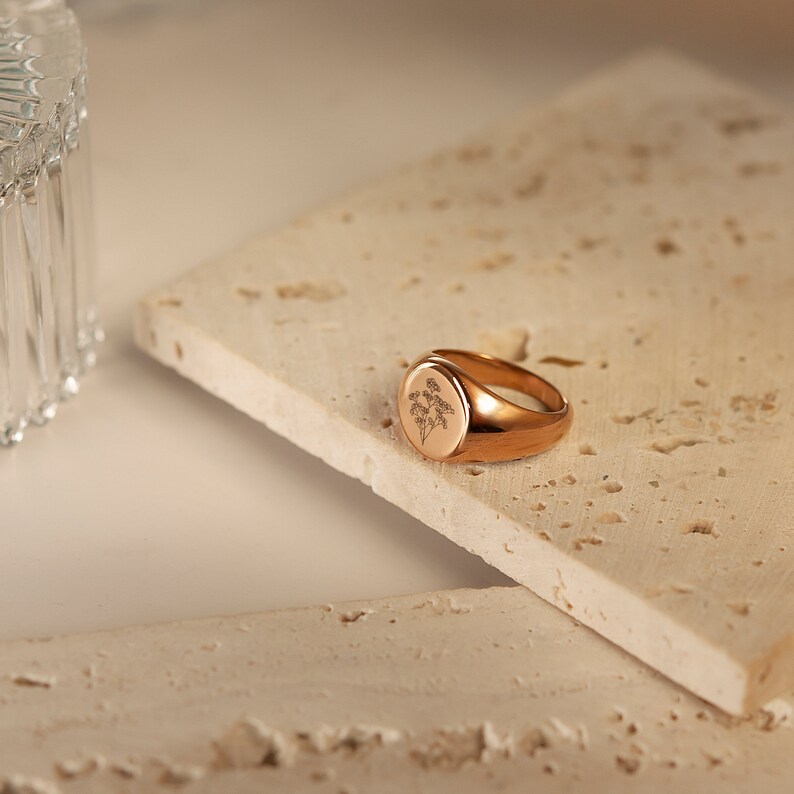May include: A gold signet ring with a floral design engraved on the top. The ring is sitting on a light brown textured surface.