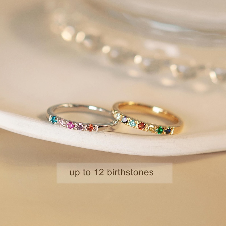 May include: Two thin rings with a row of colorful gemstones. The ring on the left is silver and the ring on the right is gold. The text "up to 12 birthstones" is below the rings.