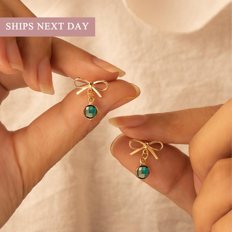 Puede incluir: Pendientes dorados con forma de lazo y una gema facetada de color verde azulado colgando. Los pendientes est&aacute;n sostenidos por los dedos de una persona. El texto "SHIPS NEXT DAY" est&aacute; en un rect&aacute;ngulo rosa en la esquina superior izquierda.