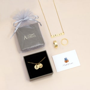 May include: An assortment of gold-colored jewelry, including a necklace, rings, and a pendant necklace with two circular charms. A gift card with the text "A gift for you" is also present, along with a gray gift box and a sheer bag.