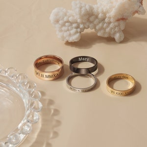 May include: Four gold, silver, and black rings with engraved text. The rings are on a light beige surface. The text on the rings reads "My Everything", "Mazy", "49.1331&deg;N, 122.8720&deg;W", "all my days", "V.II.MMXXII", and "VII.XXXI.XXII".