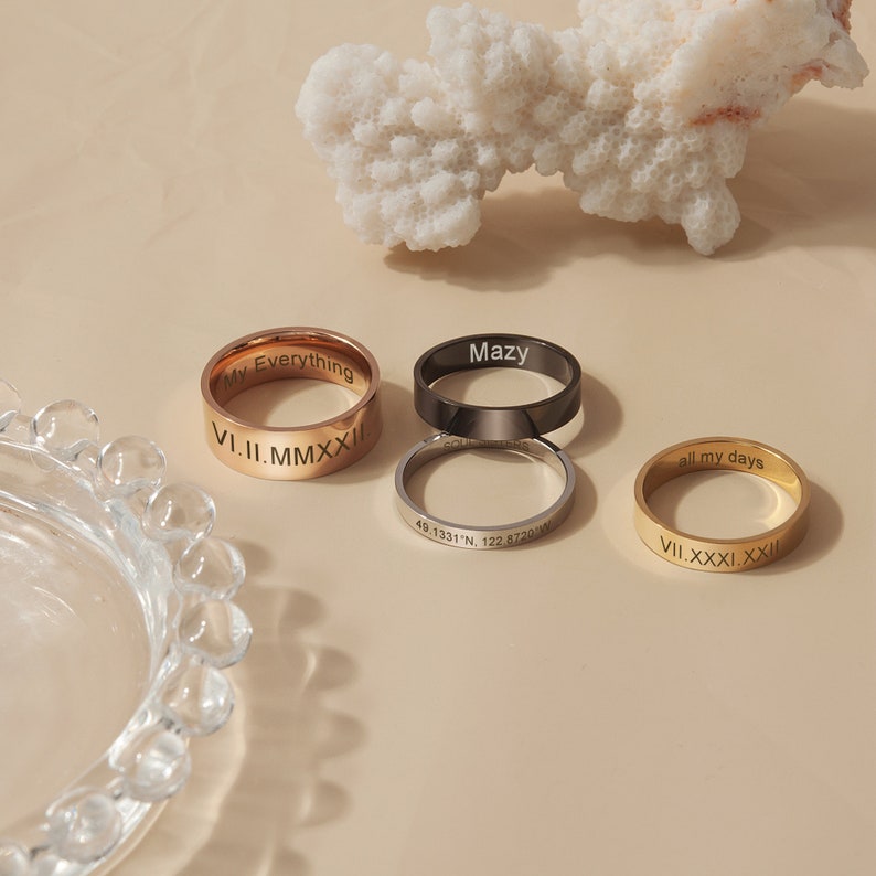 May include: Four gold, silver, and black rings with engraved text. The rings are on a light beige surface. The text on the rings reads "My Everything", "Mazy", "49.1331&deg;N, 122.8720&deg;W", "all my days", "V.II.MMXXII", and "VII.XXXI.XXII".