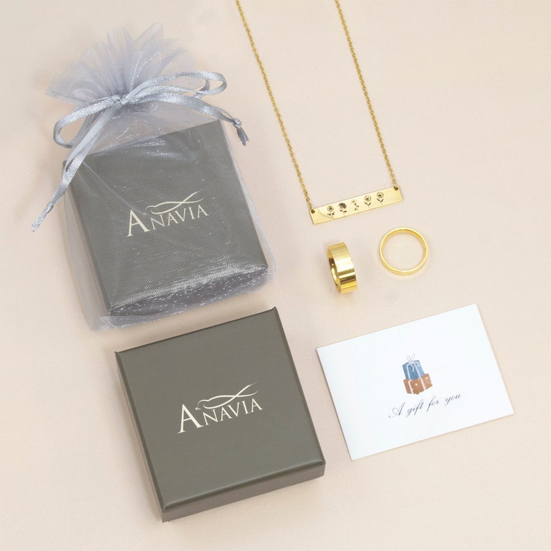 May include: A gold necklace with a bar pendant engraved with symbols. The necklace is displayed on a light pink surface with two gray boxes, a small white card with the text "A gift for you", and a gold ring.