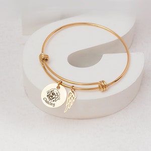 Op de afbeelding: Gouden armband met een ronde bedel met een hondenportret en de naam "Oakley". Een kleine vleugelbedel hangt naast de hondenbedel. De armband wordt gepresenteerd op een wit oppervlak.