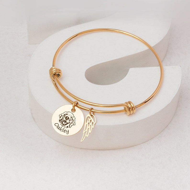 Op de afbeelding: Gouden armband met een ronde bedel met een hondenportret en de naam "Oakley". Een kleine vleugelbedel hangt naast de hondenbedel. De armband wordt gepresenteerd op een wit oppervlak.