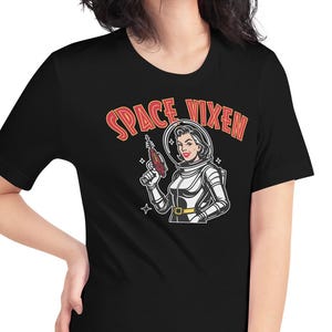 Könnte beinhalten: Schwarzes T-Shirt mit einer Retro-Illustration einer Frau in einem Raumanzug, die eine Strahlenkanone hält. Der Text "SPACE VIXEN" ist in Rot und Orange über dem Bild angeordnet. Das T-Shirt hat einen klassischen Rundhalsausschnitt.