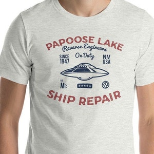 Pode incluir: Uma camiseta cinza mesclada com um gráfico vintage de um disco voador. O gráfico diz "Papoose Lake Reverse Engineers Since 1947 On Duty NV USA Mc Ship Repair".