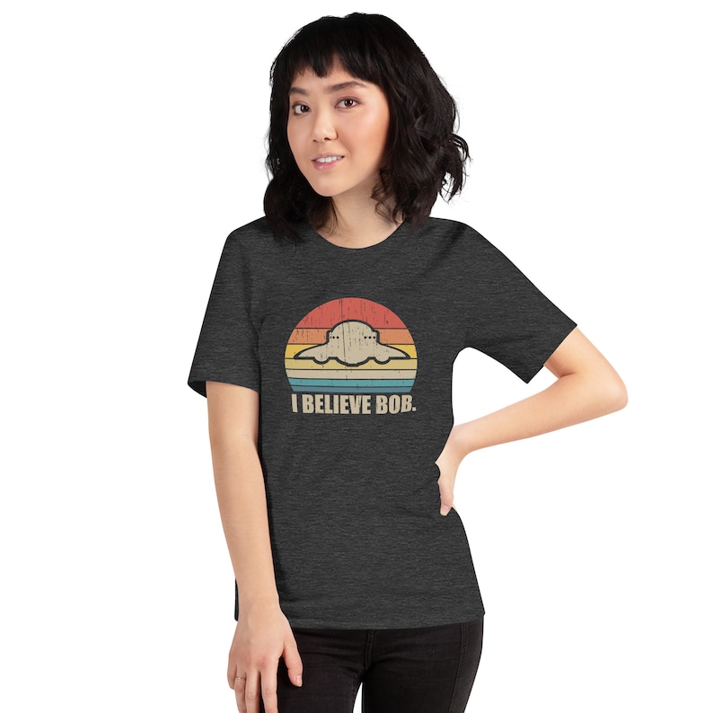 May include: A dark heather grey t-shirt with a retro-style graphic of a UFO in front of a sunset. The text "I BELIEVE BOB." is printed below the graphic.