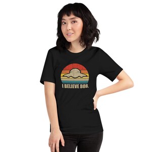 May include: A black t-shirt with a retro-style graphic of a UFO flying over a sunset. The text "I BELIEVE BOB." is printed below the graphic.