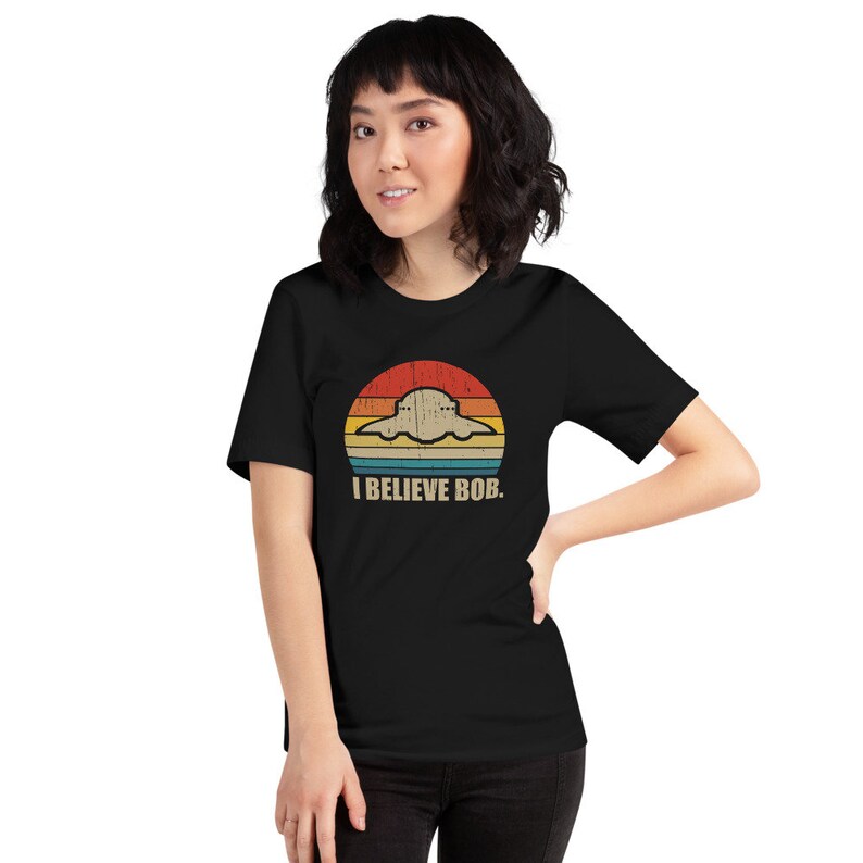 May include: A black t-shirt with a retro-style graphic of a UFO flying over a sunset. The text "I BELIEVE BOB." is printed below the graphic.