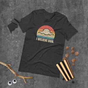 May include: A dark heather grey t-shirt with a retro-style graphic of a UFO with the text "I believe Bob."