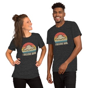 May include: Two people wearing dark grey t-shirts with a retro sunset graphic and the text "I Believe Bob." The graphic features a cartoon ghost in a flying saucer.