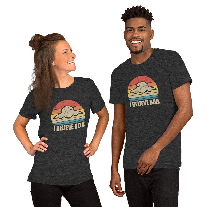 May include: Two people wearing dark grey t-shirts with a retro sunset graphic and the text "I Believe Bob." The graphic features a cartoon ghost in a flying saucer.